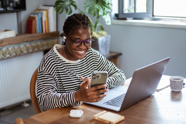 Woman sits at her desk with a laptop while smiling at her smartphone.