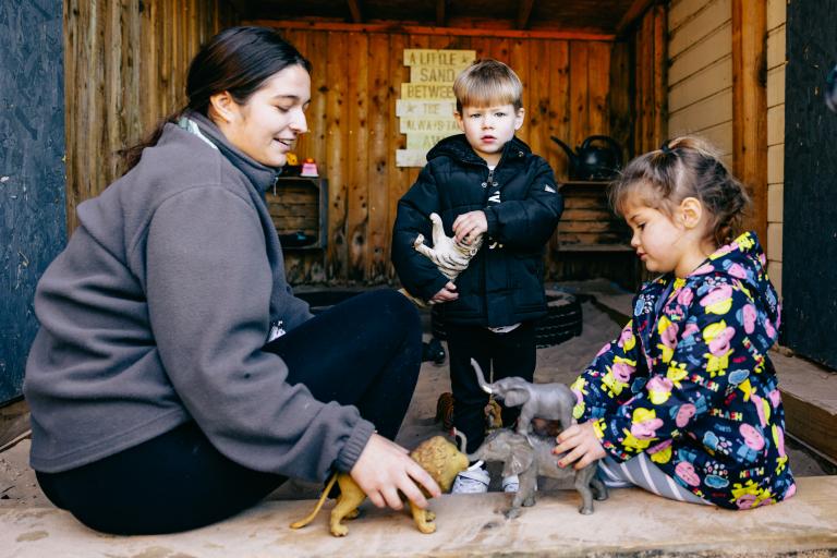 Early years practitioner sat outside with two young children playing with toy animals.