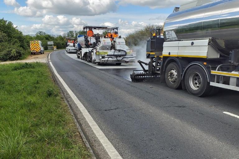 Two large resurfacing vehicles laying new surface on a stretch of road on a sunny day. 