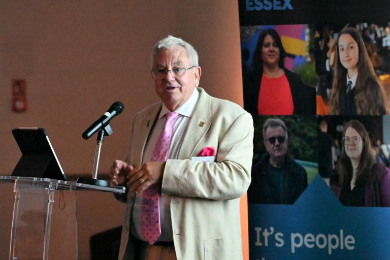 Cllr Durham gives a speech at a podium in a beige suit in front of an Extraordinary Essex banner.