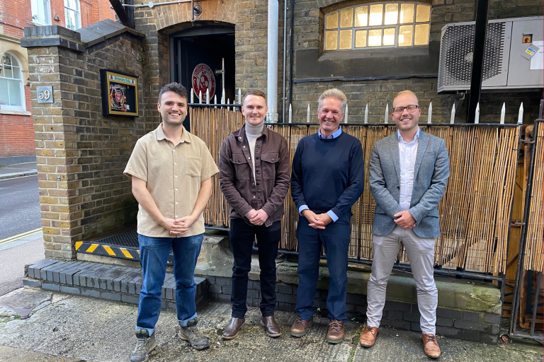 Four men smiling stood in front of a brick building.
