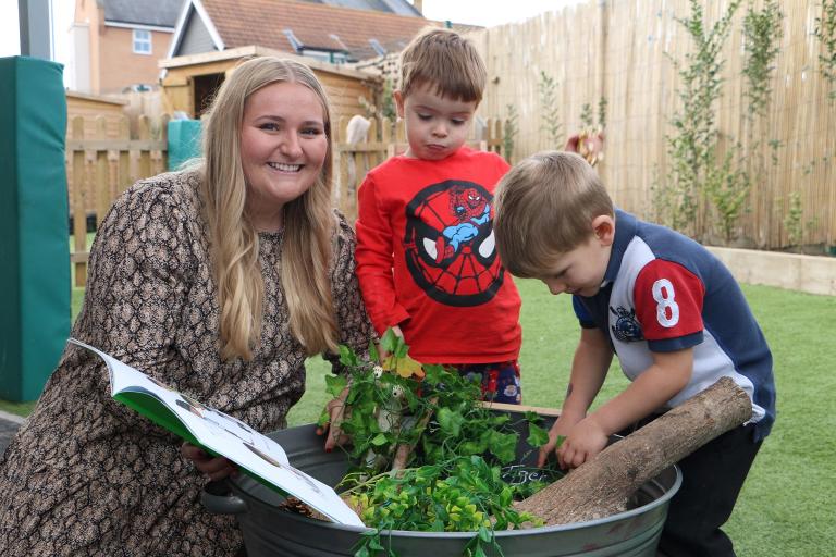 A woman reads to two little boys outside as they play with a plant pot.