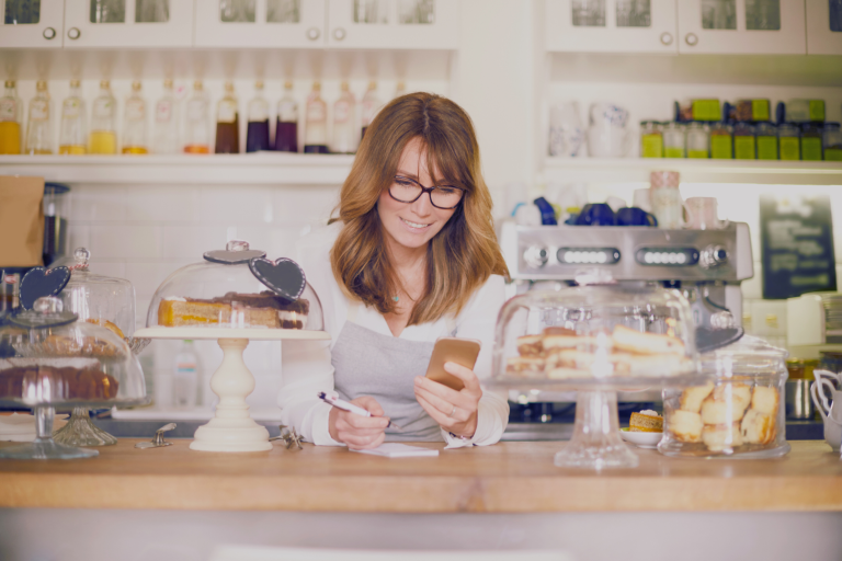 Woman sitting behind her cafe, working on her phone and smiling