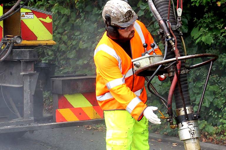 A worker dressed in high-visibility jacket and hard hat spreading asphalt on a road surface using machinery.