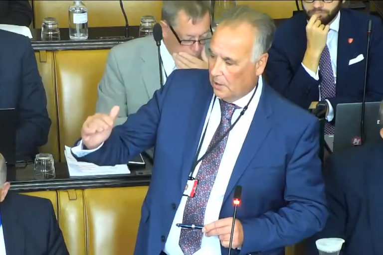 Men in suits sit in the council chamber listening to the chancellor speak.
