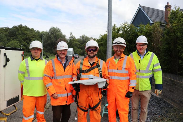 Five men wearing high-visibility jackets and hard hats holding an LED streetlight.