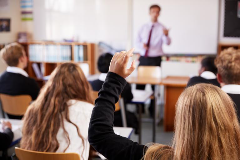 From the back of a classroom, a child raises her hand to the teacher out of focus.