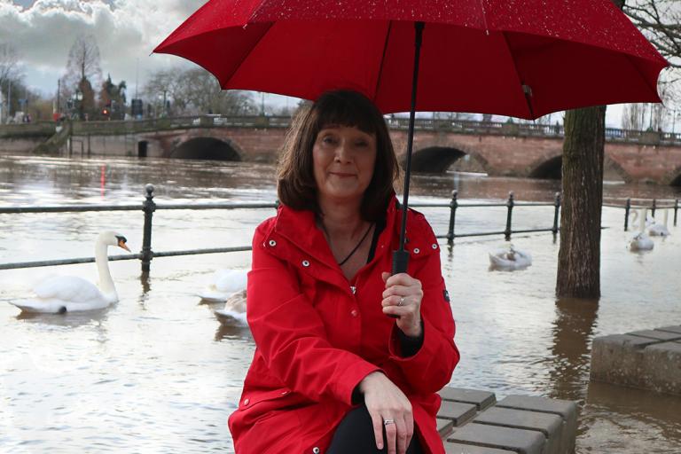 Lady in a red raincoat with a red umbrella sits in front of an overflowing river.