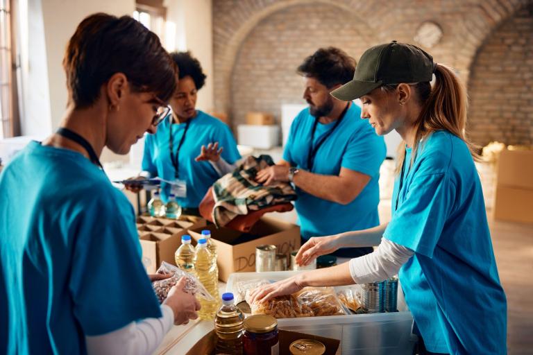 Four volunteers wearing blue t-shirts stand packing food into boxes in a hall.