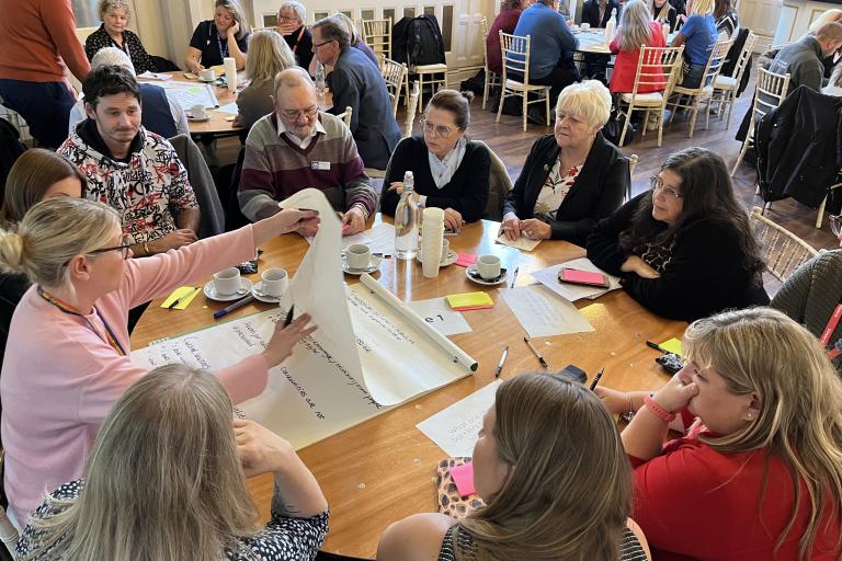 Groups of people sit at several roundtables while discussing and mindmapping ideas onto large sheets of paper.