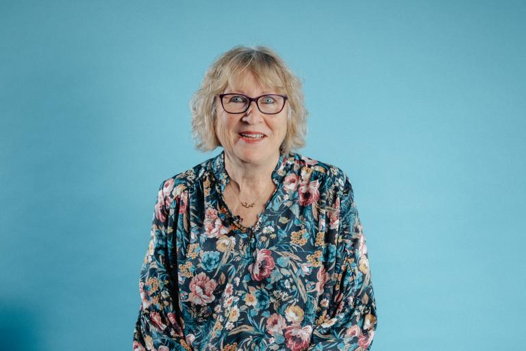 Headshot image of Karen, a white woman with short white hair, a fringe and glasses. She is sitting on a chair in front of a blue studio background.