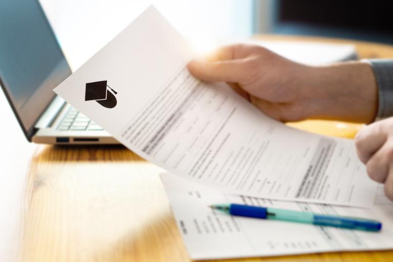 Close-up of man's hands reading school application paper on desk with laptop in background.