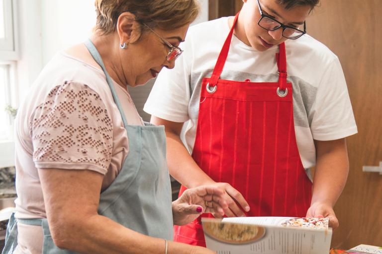 An older woman and a teenager looking at a cookbook. They're both wearing aprons and standing at a kitchen worktop.