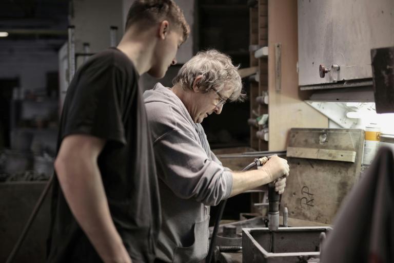An older man and a teenage boy standing in a garage, it's quite dark and industrial. There is an open toolbox on the side, suggesting they are building or mending something. They are looking down at a workbench.
