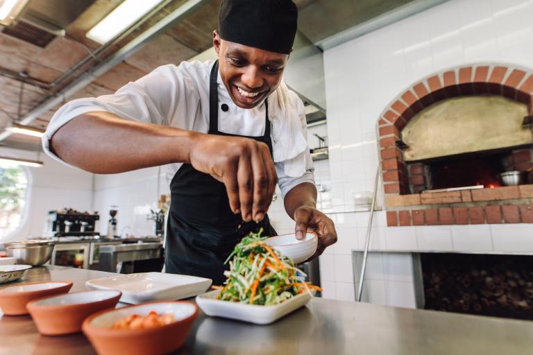 Young man in a commercial kitchen setting, wearing chef whites. He's smiling and leaning forward to garnish a dish on the kitchen counter.