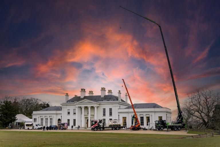 A photograph of a grand house with large cranes outside the front. A red and blue sky is in the background. 
