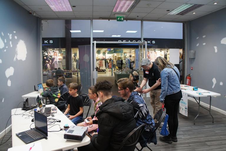 A group of young people sitting at a desk inside a shop with a large windowfront. 
