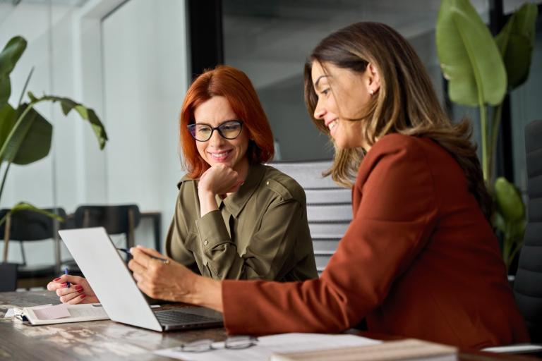 Two women sitting at a desk in an office. They are both smiling while looking at a laptop screen and holding pens in their hands.