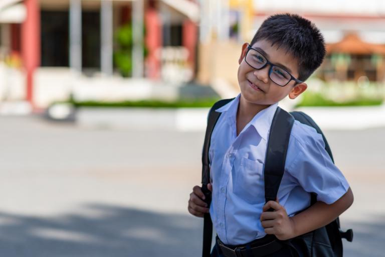 Young boy (primary school age) wearing a white school shirt, glasses and a khaki backpack. He is smiling and tilting his head to one side.