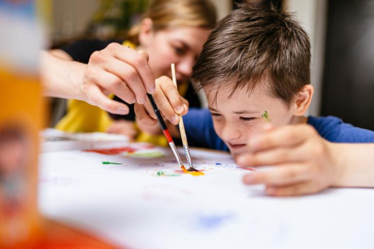 A female teacher and young male pupil sitting and painting together.