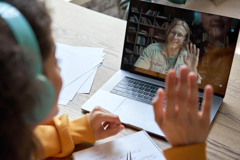 Over-the-shoulder shot of a school girl on a video call through her laptop to a man waving back.