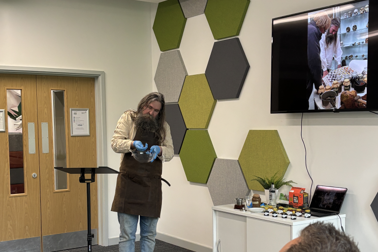 A man with a large beard wearing gloves and an apron standing at the front of a room with a TV screen on the wall.