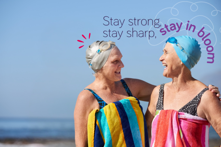 Two ladies aged 60 plus in their swimming costumes smiling at each other after a swim in the sea. The text says 'stay strong, stay sharp, stay in bloom'.
