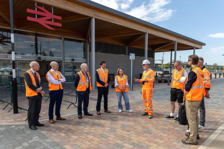 Group of men and women in hi-vis outside terminal of new train station listening to developers.