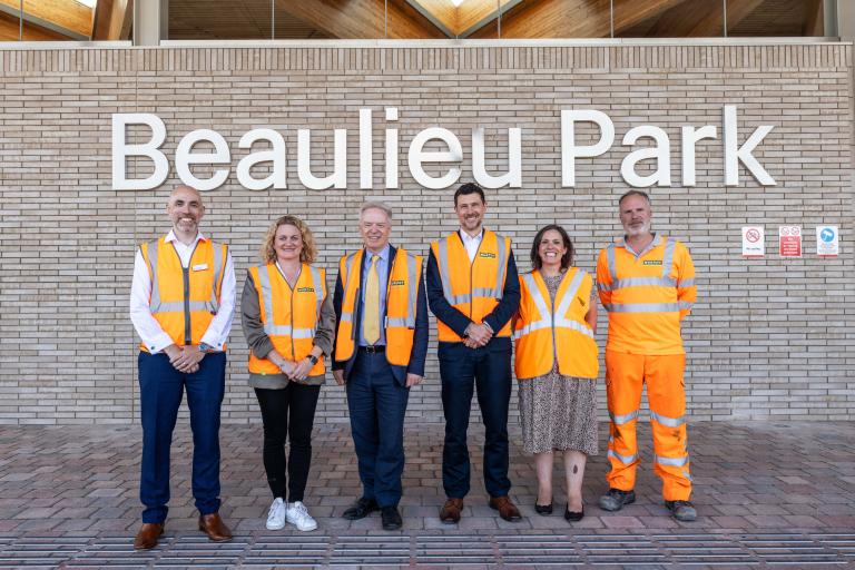 Group of men and women in hi-vis face camera in front of brick wall with train station terminal in background.