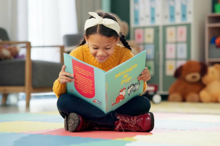 Young girl reading book while cross-legged on floor surrounded by toys and stuffed animals.