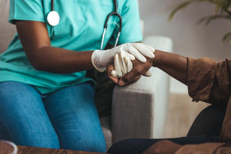 A shot of a female nurse wearing a glove holding the hand of an elderly gentleman. The nurse has a stethoscope around her neck.