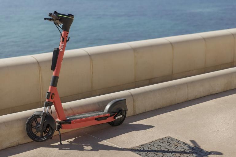 An orange electronic scooter leaning up against a wall. Behind is the sea.