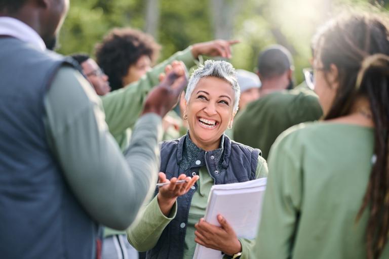 A woman smiling facing the camera holding a clipboard. She is in the middle of a group of people. They are all wearing similar clothes.
