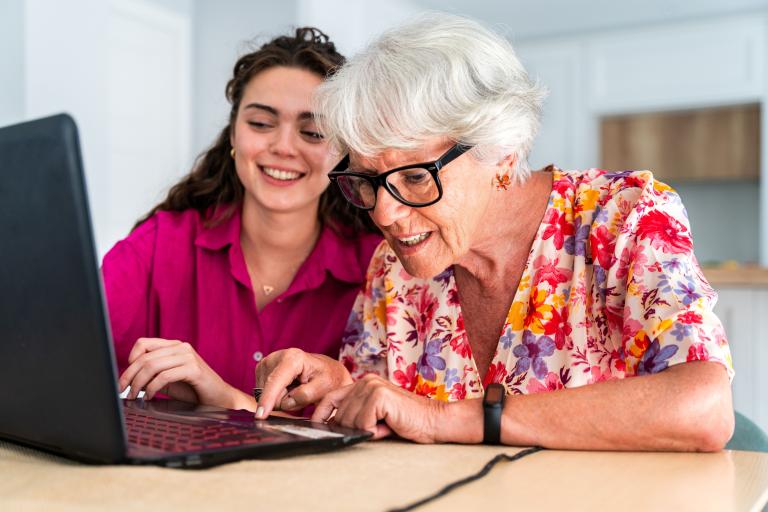 Two people sitting down, one helping the other on a laptop.
