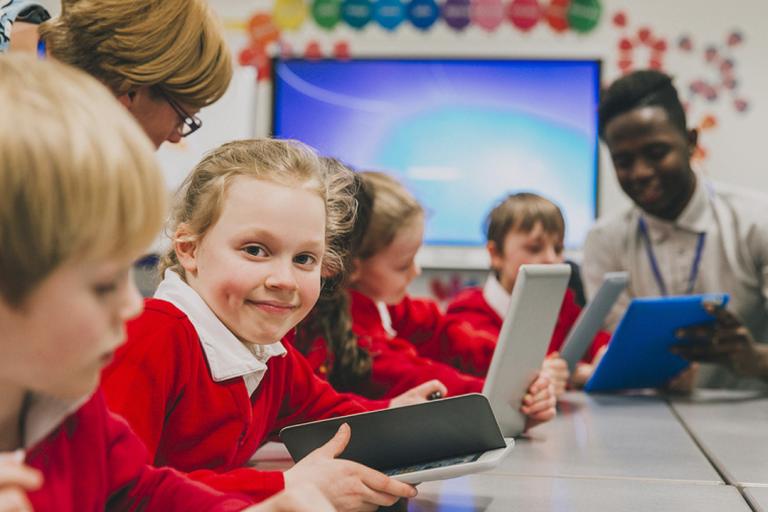 Primary school children sat at a desk looking at digital tablets. One girl is smiling at the camera.