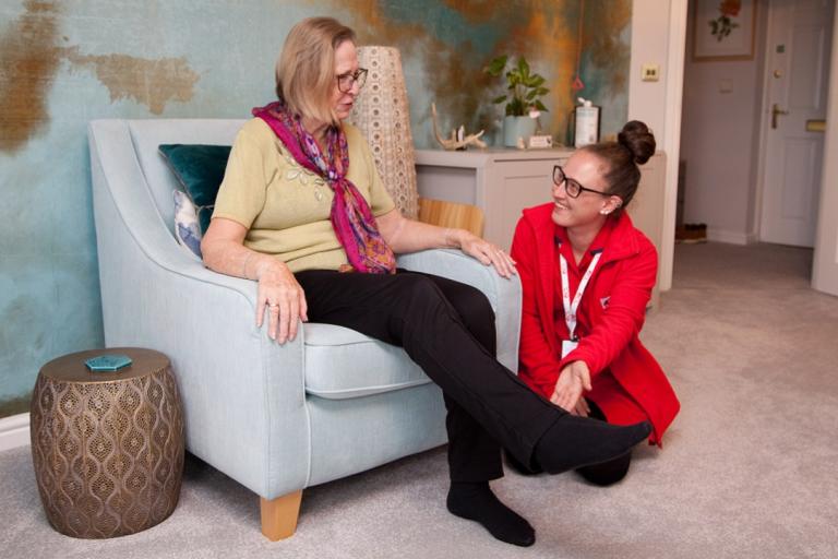A carer in a red uniform crouches down and supports the ankle of an older woman in an armchair.