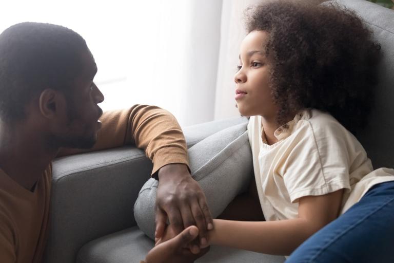 A father holds his daughter's hand while she lies on the sofa looking upset.