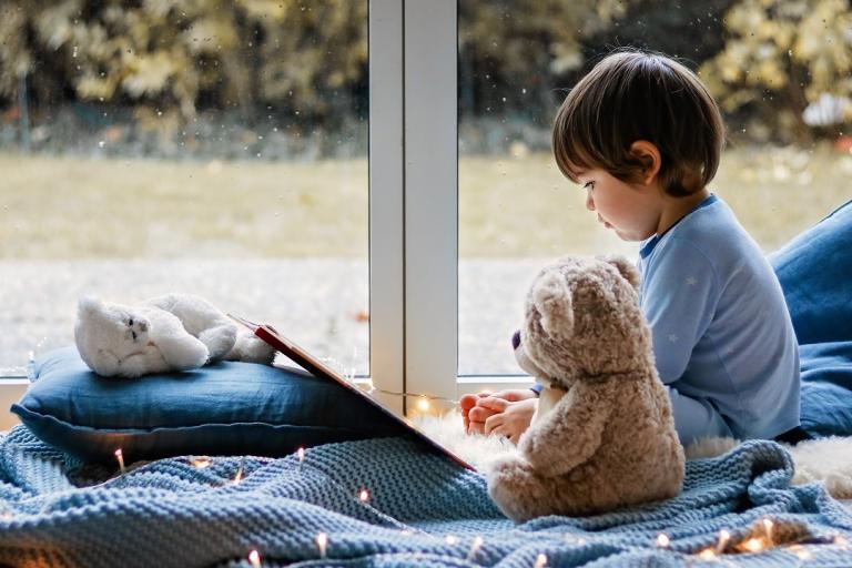 A little boy reading book with his teddy bear toy sitting cozy on pillows and knitted blanket near wet window with autumn garden at background.