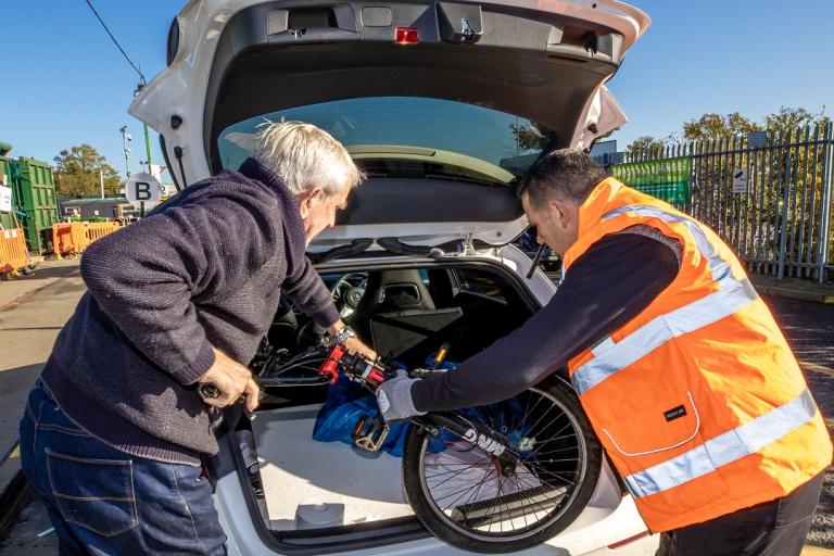 A recycling centre staff operative helps a gentleman lift a bike from the boot of a vehicle. 