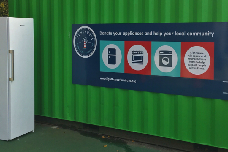 A green recycling container with a Lighthouse donation point sign and an upright fridge.