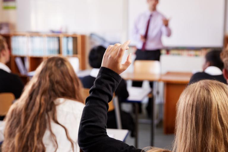 A female pupil raising her hand in a classroom, with the teacher at the front of the class.