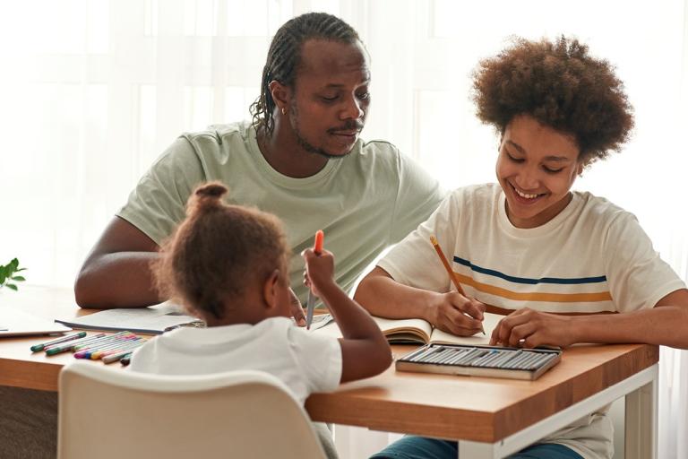 A man sits at a dining table helping his child with their schoolwork.