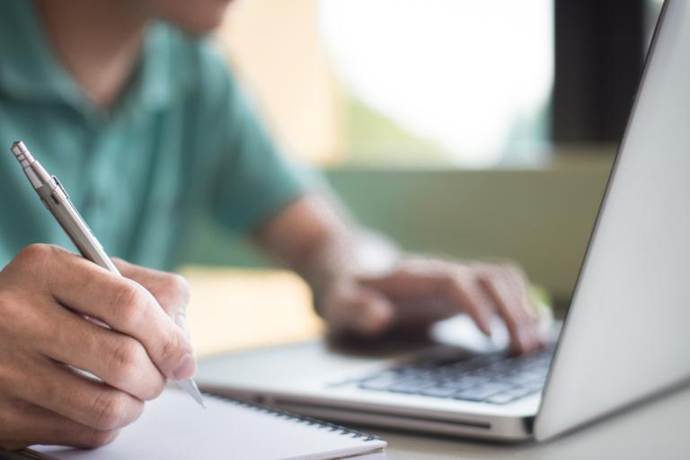 A man with a notepad and pen looking at a laptop