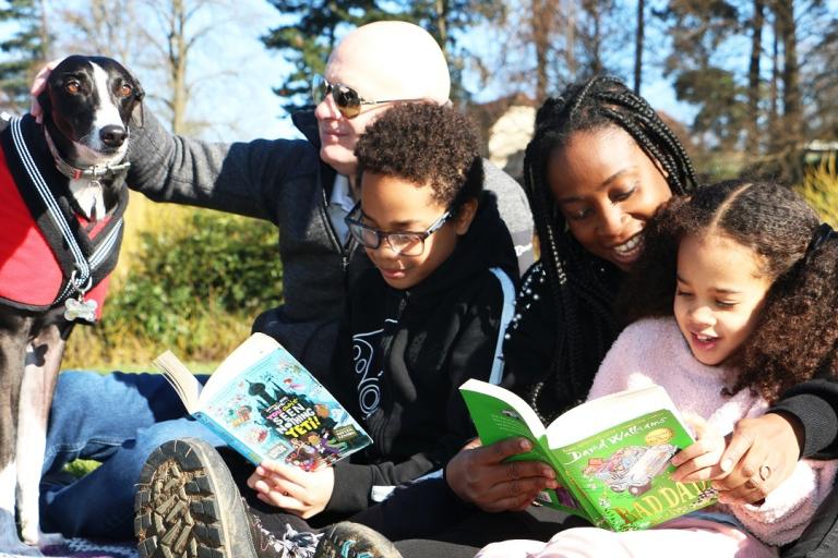 A mother and father sit reading books with their two children and dog in a park.