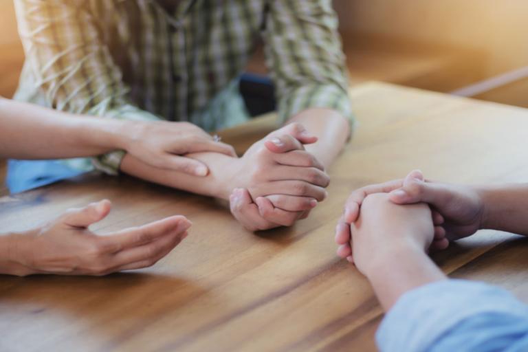 A close up of three people sitting around a table with their hands together. One person is reaching out and touching of the other person's arms.