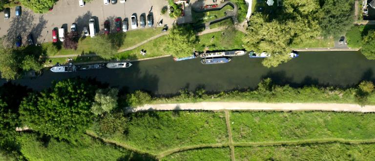 An aerial view of a river in Harlow. There are several barges stationary in the river. On each side of the river there are fields and trees, and footpaths which follow the river. On one side of the river there is a car park with several cars parked.